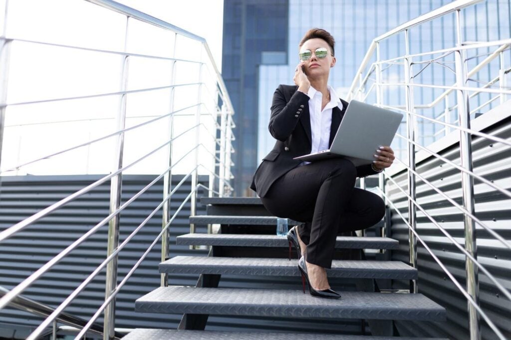 A businesswoman with a laptop in sunglasses sits on top of a staircase, busy with work.