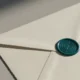 Close-up of a deep-teal wax seal with an abstract geometric emblem on a blank envelope, set on a soft neutral background.