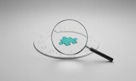 Magnifying glass spotlighting a unique teal puzzle piece fitting into a neutral circular puzzle on a soft gray background.