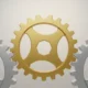 Golden uniquely shaped gear meshing between two silver gears on a soft neutral background.